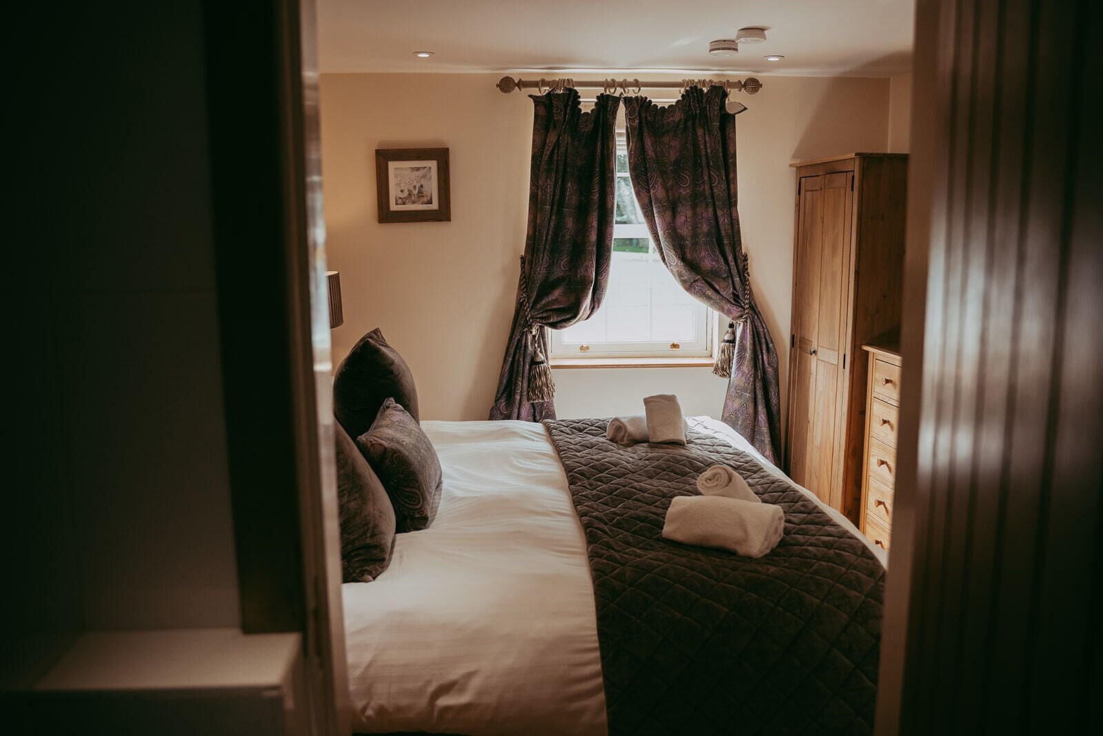 A bedroom with a bed, pillows, a quilted blanket, a wooden wardrobe, and a window with curtains.