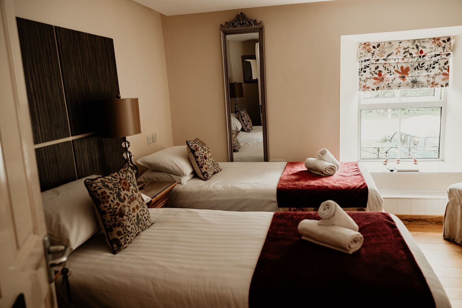 A hotel room with two beds, a large mirror, a window with floral curtains, and a view of the outside.