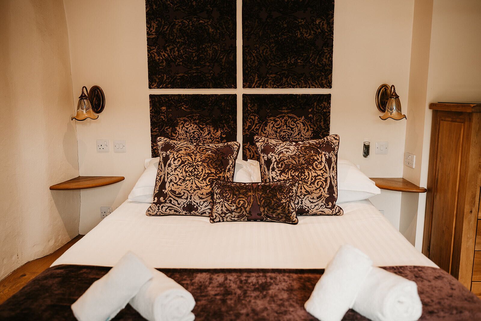 Habitación de hotel con una cama grande, almohadas decorativas, cabecera de madera, mesitas de noche y un armario de madera.