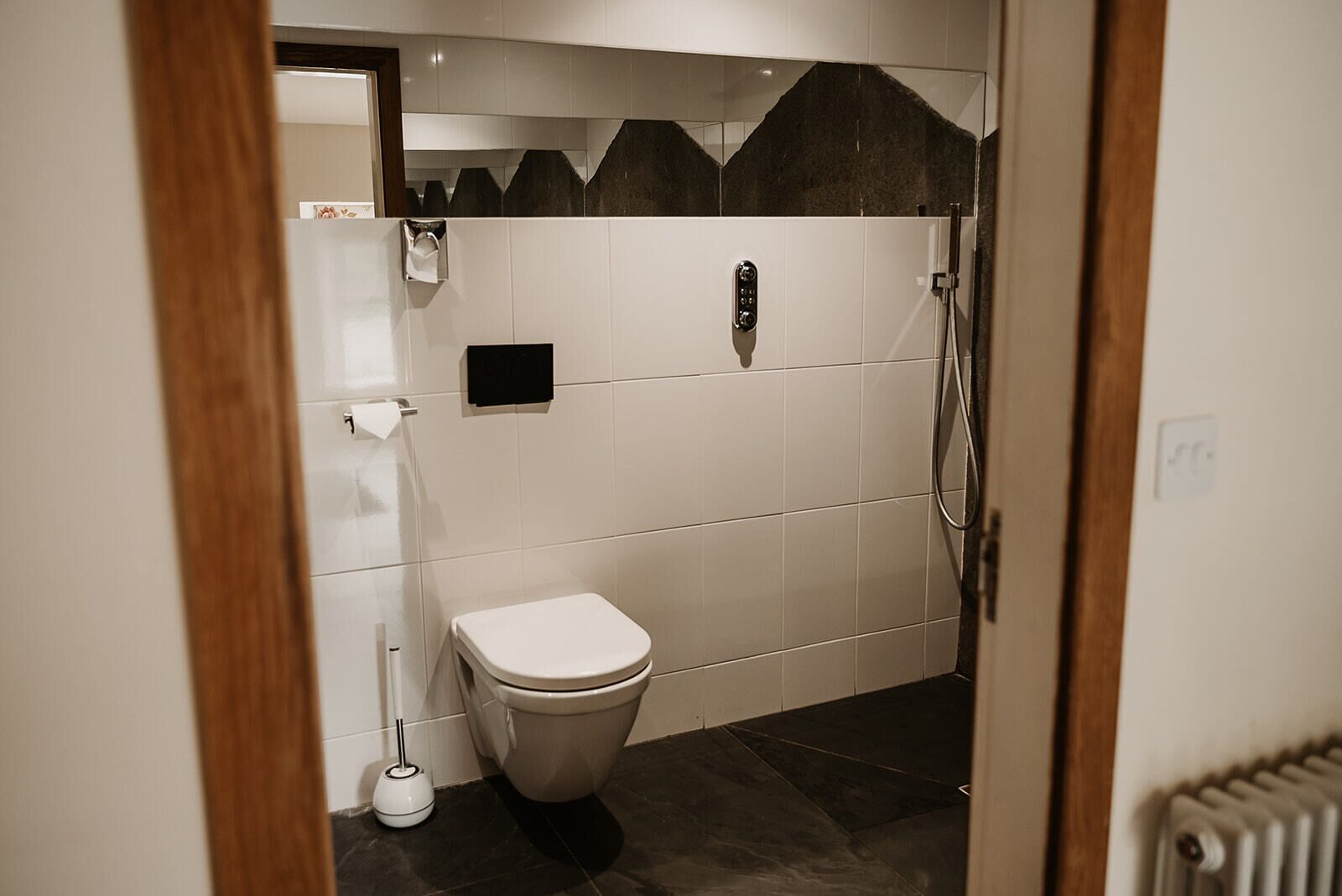 A small, modern bathroom with white tiles, a toilet, a shower, and a towel dispenser.