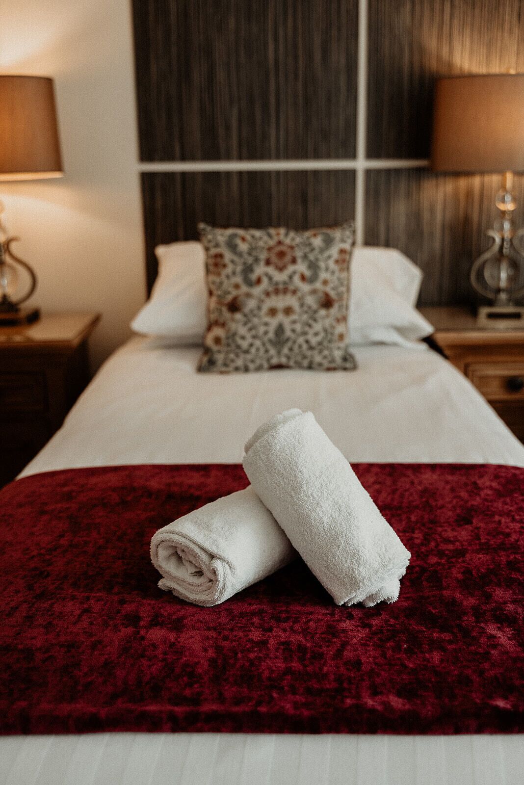Un lit bien fait, recouvert d’une couverture rouge, avec deux oreillers blancs et un coussin décoratif. On y trouve deux lampes de chevet et une petite table avec une lampe.