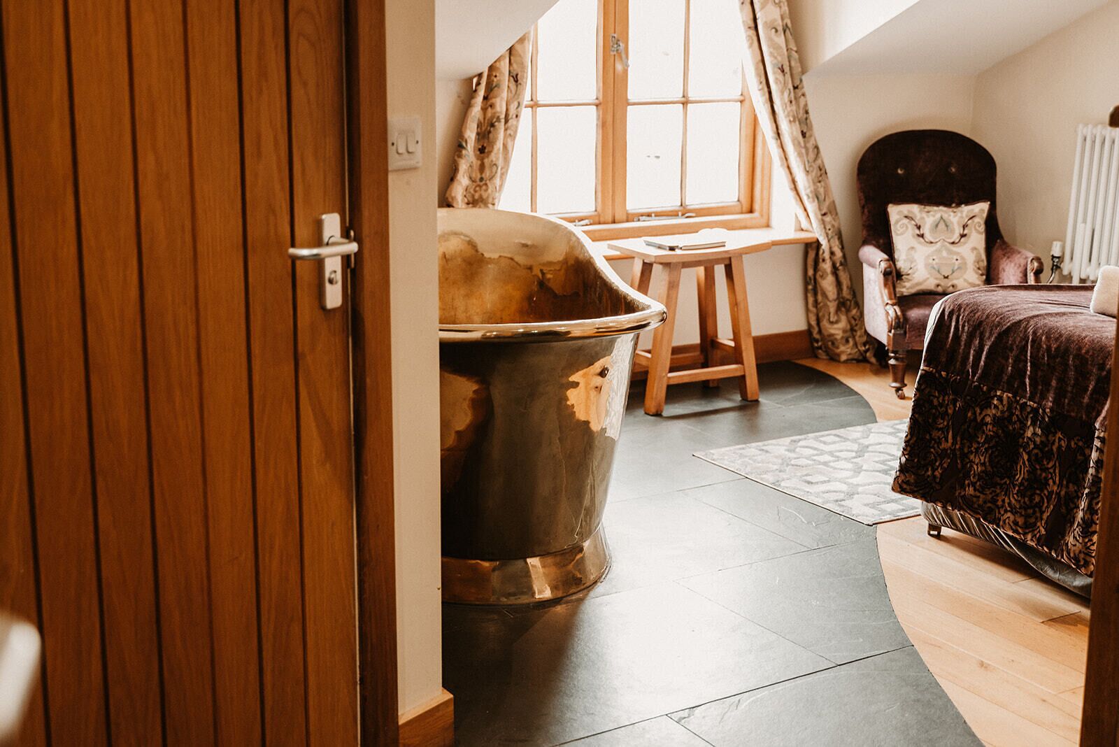 Una habitación con una bañera grande, una puerta de madera, una ventana con cortinas, una cama y una silla.