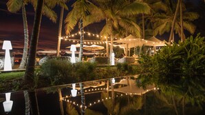 A nighttime outdoor area with string lights, palm trees, and a tented seating area by a reflective water surface. - Ness By D-Ocean (Saint-Paul)