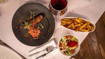 Una mesa con un plato de comida, un bol de ensalada, una copa de vino tinto y una cesta de papas fritas.