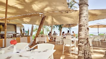 An outdoor dining area with tables and chairs under large umbrellas, overlooking a beach.