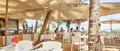 An outdoor dining area with tables and chairs under large umbrellas, overlooking a beach.