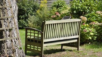 A wooden bench with slatted backrest and armrests, situated in a garden with various plants and a wooden fence in the background.