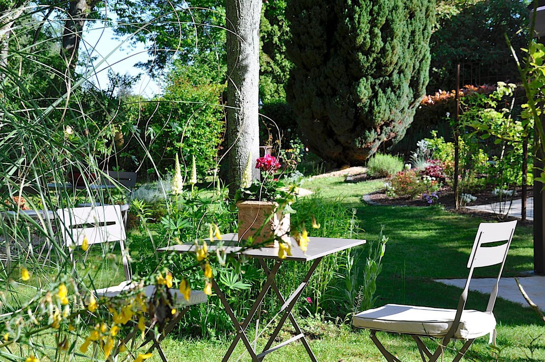 Un jardín al aire libre con una mesa y dos sillas, rodeado de diversas plantas y un árbol grande.