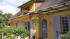 A yellow building with a brown roof, white shutters, and a covered porch with hanging flower baskets. - Au Jardin de Lisa (Cherré-Au)