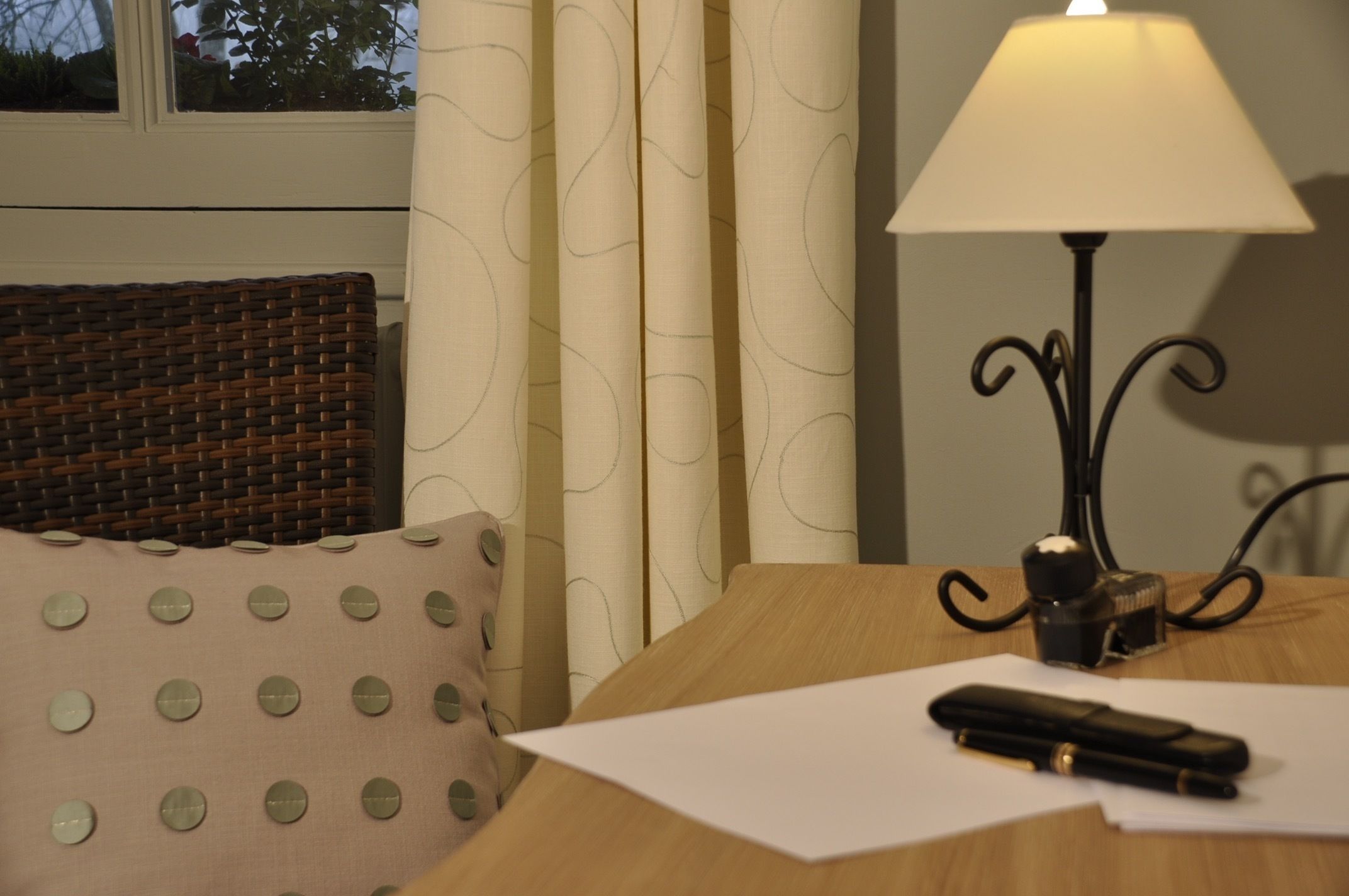 Una habitación con cabecera de mimbre, un cojín con lunares, una lámpara y un bolígrafo sobre una mesa.
