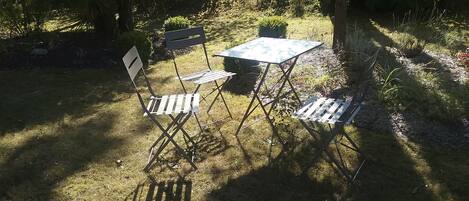An outdoor seating area with a table and two chairs, surrounded by greenery and trees.