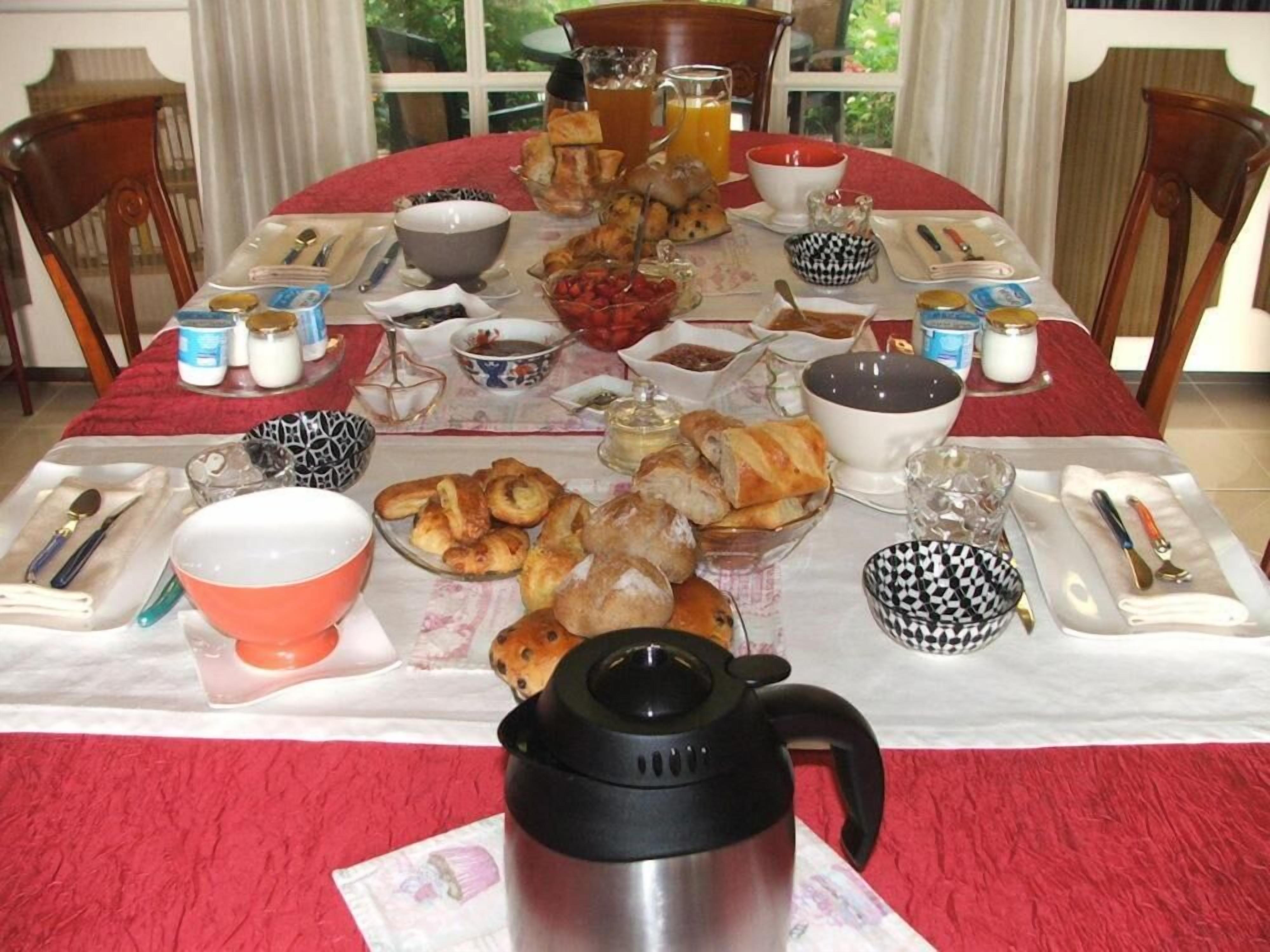 Una mesa de desayuno servida con diversos platos, entre ellos pasteles, frutas y bebidas, en una habitación con vista a la vegetación.