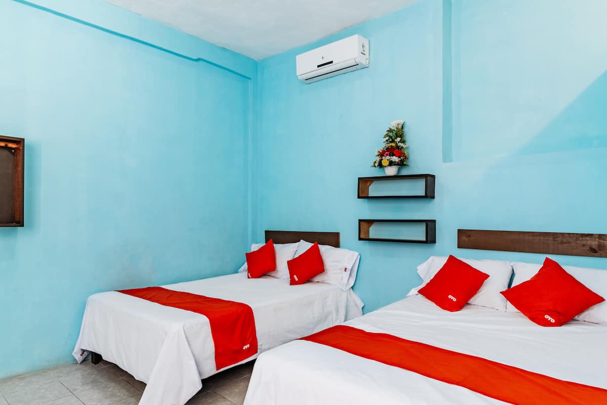 A hotel room with two beds, each with white bedding and red accents, a small shelf with a floral arrangement, and a wall-mounted air conditioner.