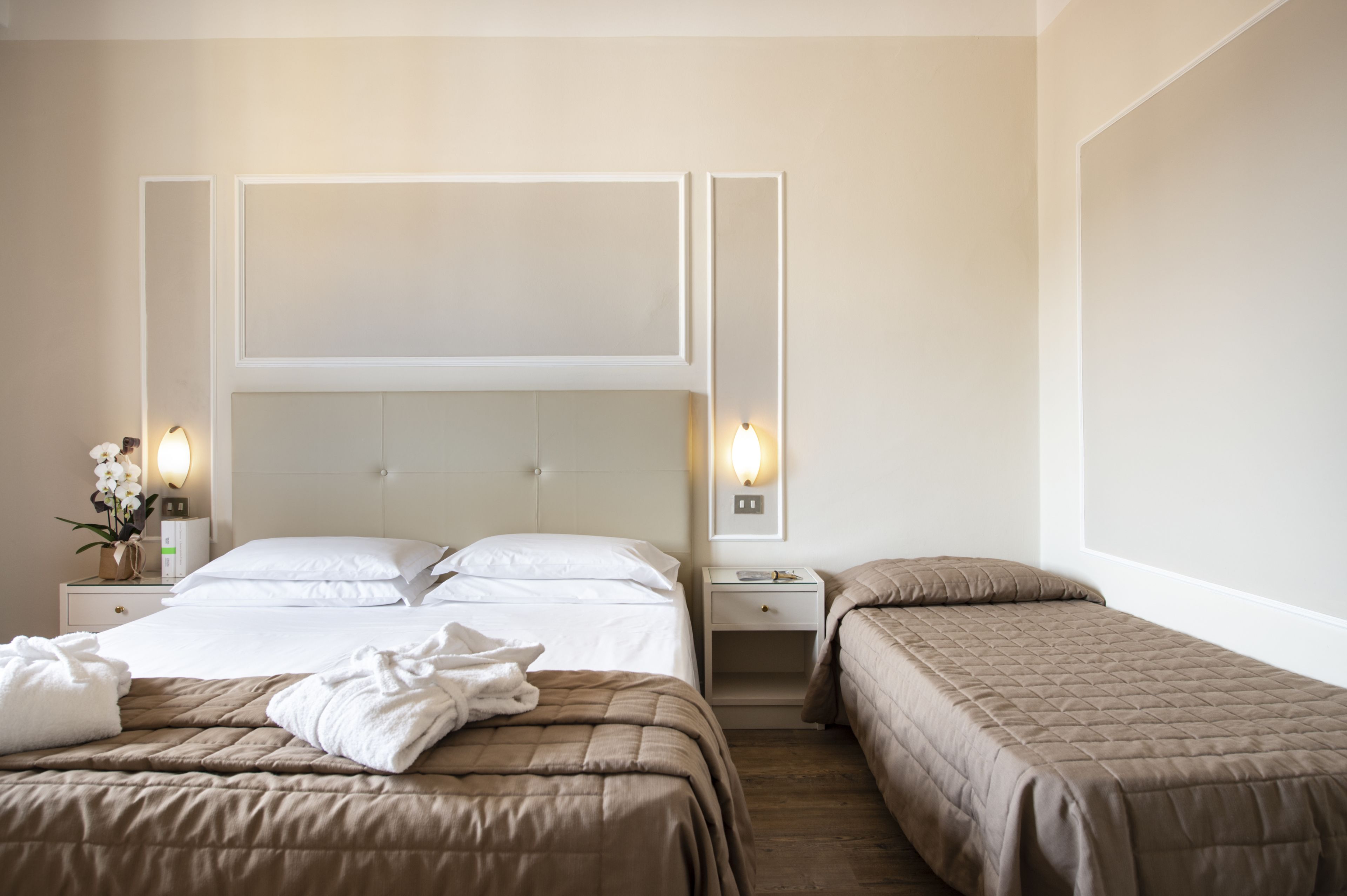 A hotel room with two beds, a nightstand with a lamp, and a wall-mounted light fixture.