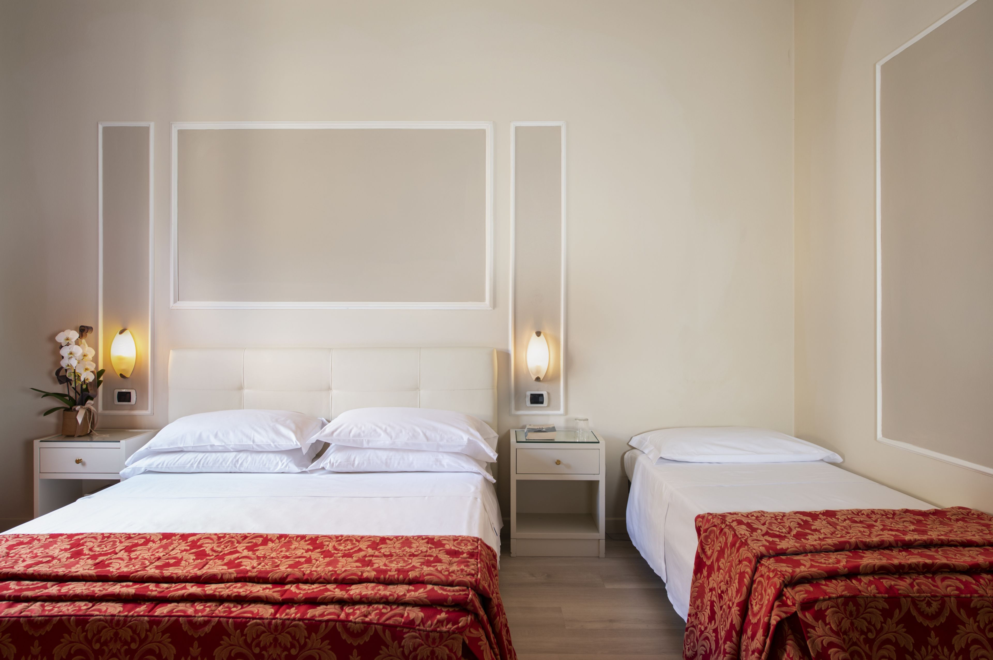 Habitación de hotel con dos camas, cada una con ropa blanca de cama y colchas estampadas en rojo. Hay mesitas de noche con lámparas y un aplique en la pared sobre las camas.