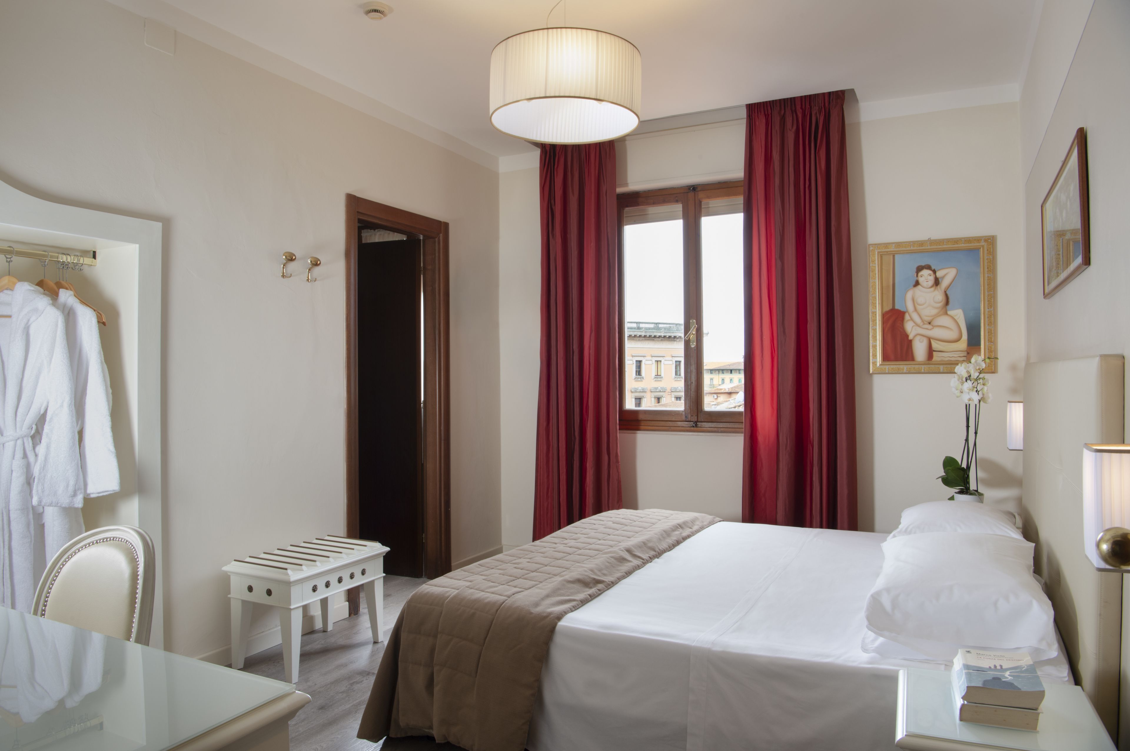 A hotel room with a bed, a bedside table with books, a mirror, a window with red curtains, and a view of buildings outside.