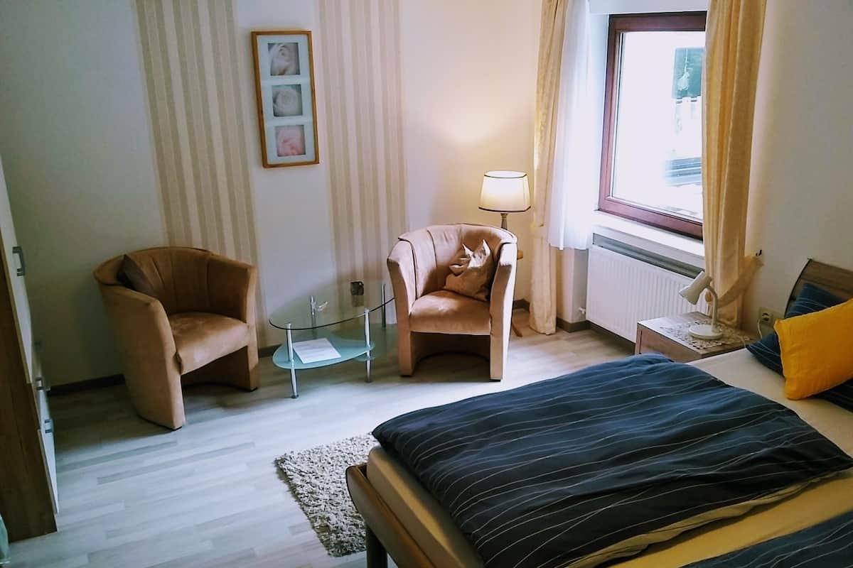 A hotel room with a bed, two armchairs, a glass coffee table, a lamp, and a window with curtains.