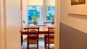 Ein Essbereich mit einem für vier Personen gedeckten Tisch, zwei Stühlen, einem Blumentopf und einem Fenster mit Blick auf ein Gebäude.