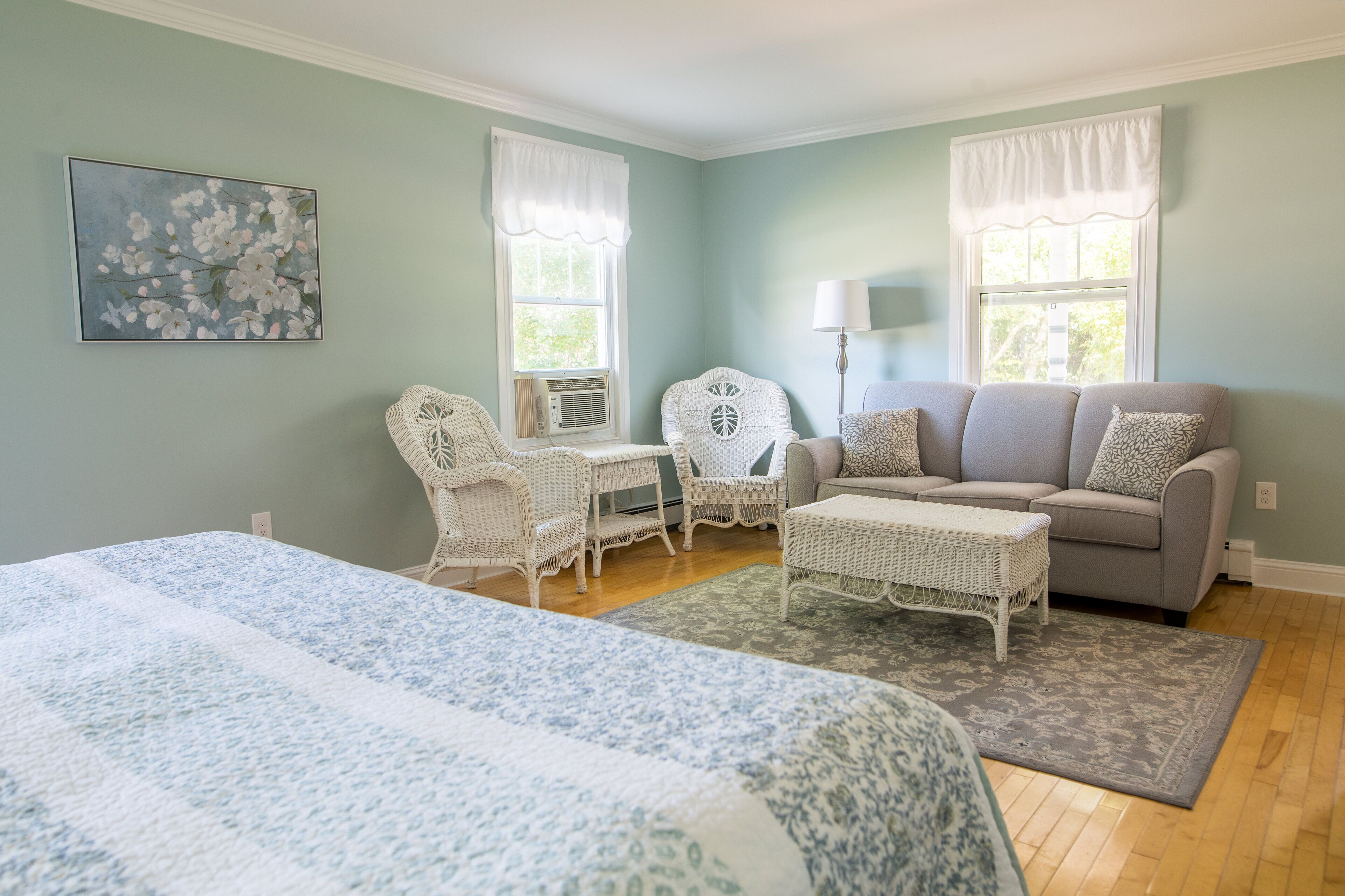A bedroom with a bed, a gray sofa, two white wicker chairs, a small table, and a lamp.