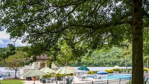 Zona de piscina con sillas de descanso y sombrillas, rodeada por una valla y árboles.