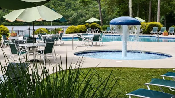 Zona de piscina con sillas de descanso y sombrillas, rodeada de vegetación y árboles.