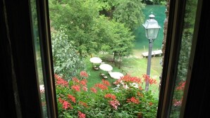 Blick durch ein Fenster auf einen üppigen Garten mit Teich, umgeben von Bäumen und roten Blüten.