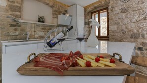 Ein Holzbrett mit Wurstwaren, Käse und Tomaten in einer Küche mit Steinwänden, einer Weinflasche und Gläsern.