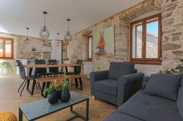 A spacious living area with a stone wall, a dining table with chairs, and a gray sofa. - Vila Magdalena (Supetar)