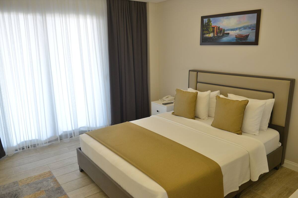 Quarto de hotel com cama, mesa de cabeceira, um quadro e janela com cortinas.