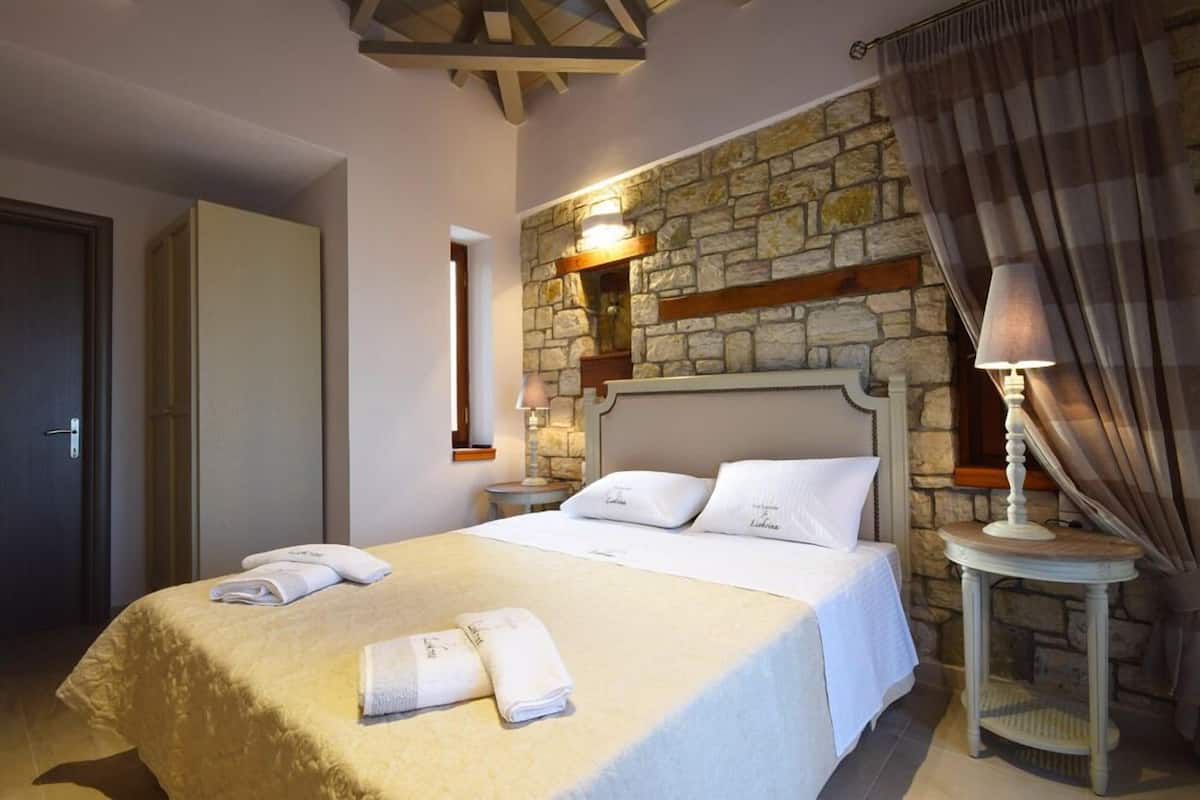 Un dormitorio con pared de piedra, una cama con ropa blanca, una mesita de noche con lámpara y una puerta.