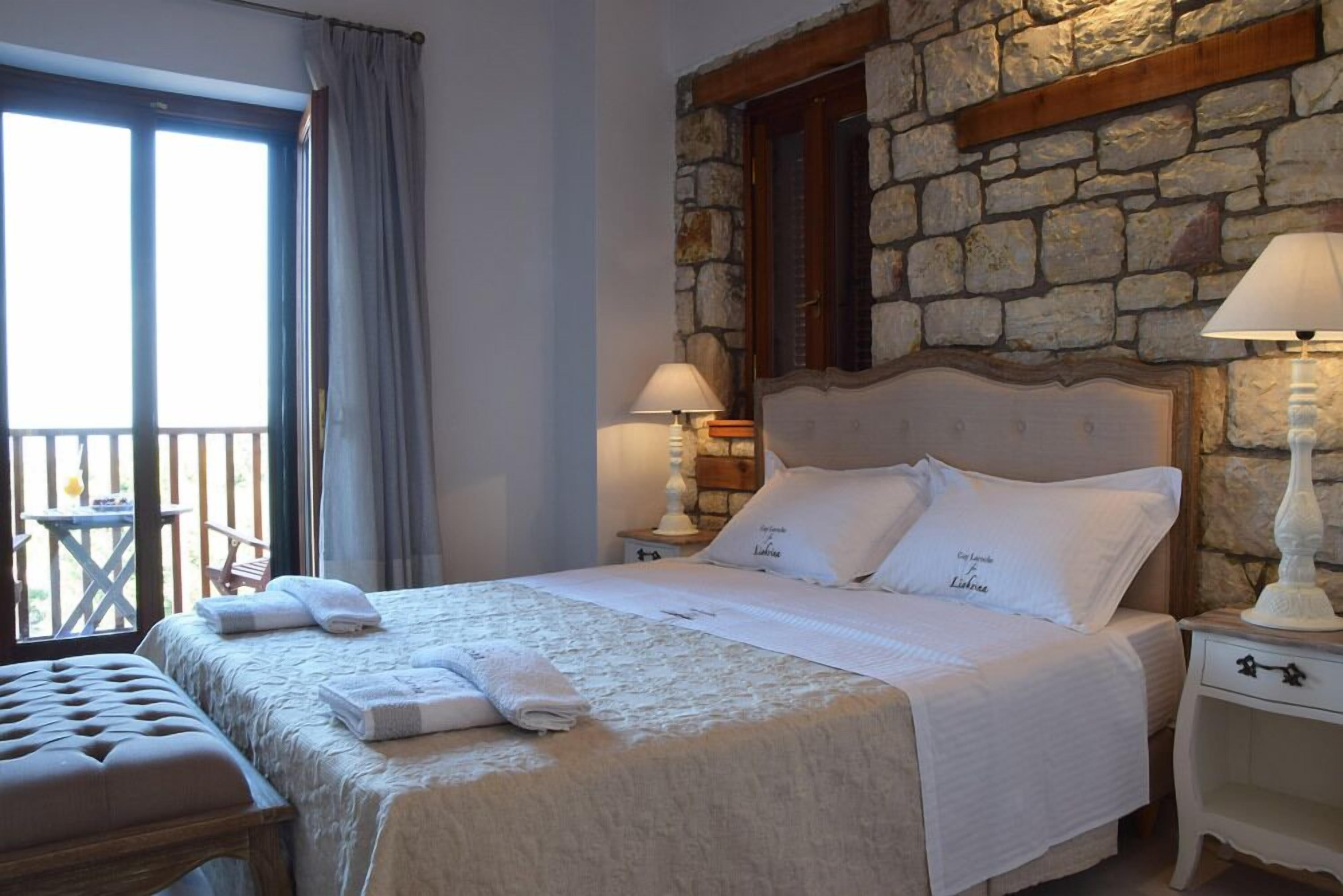 A neatly made bed with white linens, a stone wall, and a balcony with a table and chairs.