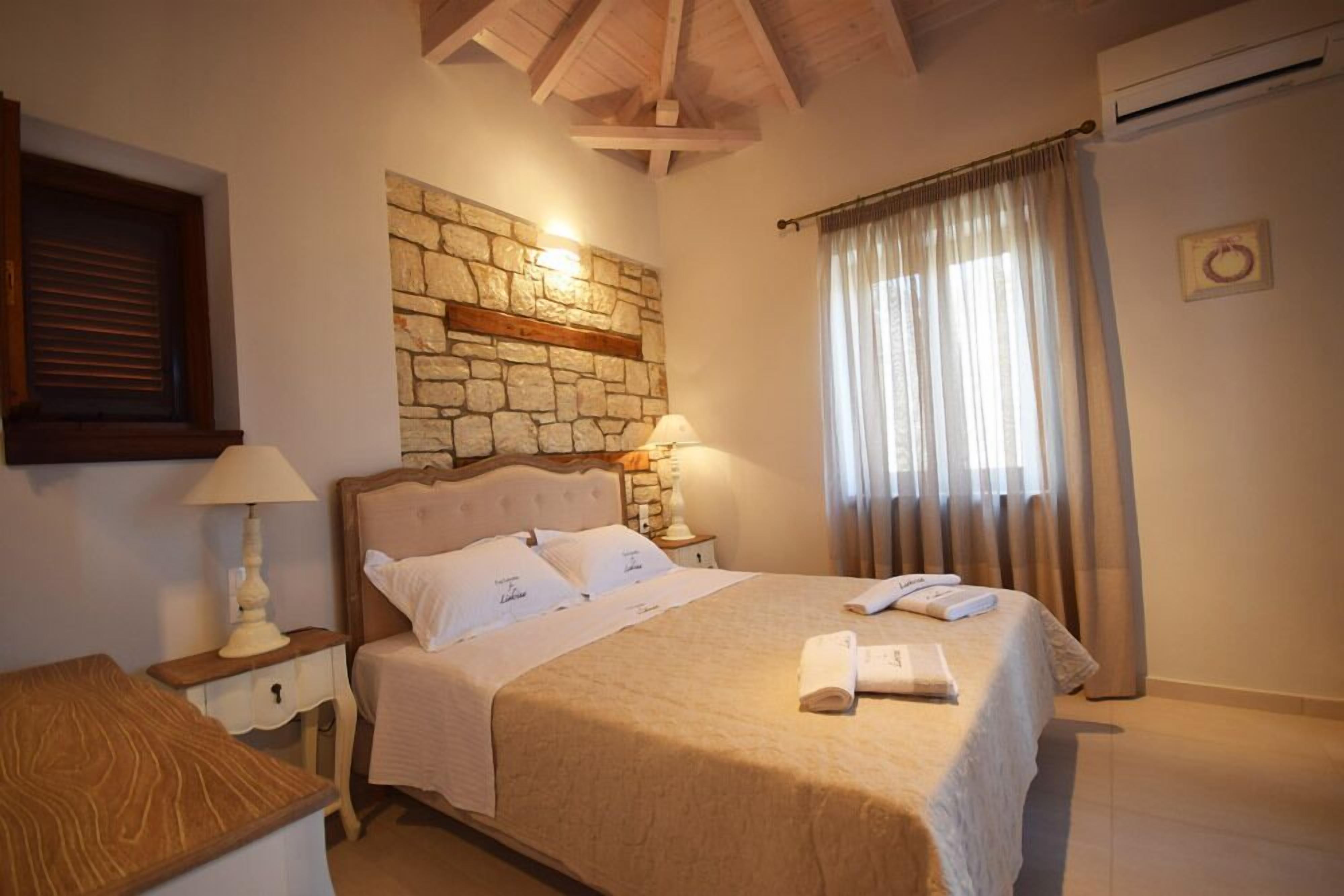 A bedroom with a stone wall, a bed with white linens, a nightstand with a lamp, and a window with curtains.
