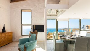 A room with a large window, a television, a wooden cabinet, a blue armchair, and a dining area with a table set for two. - Yellow Dream Villas (Chania)