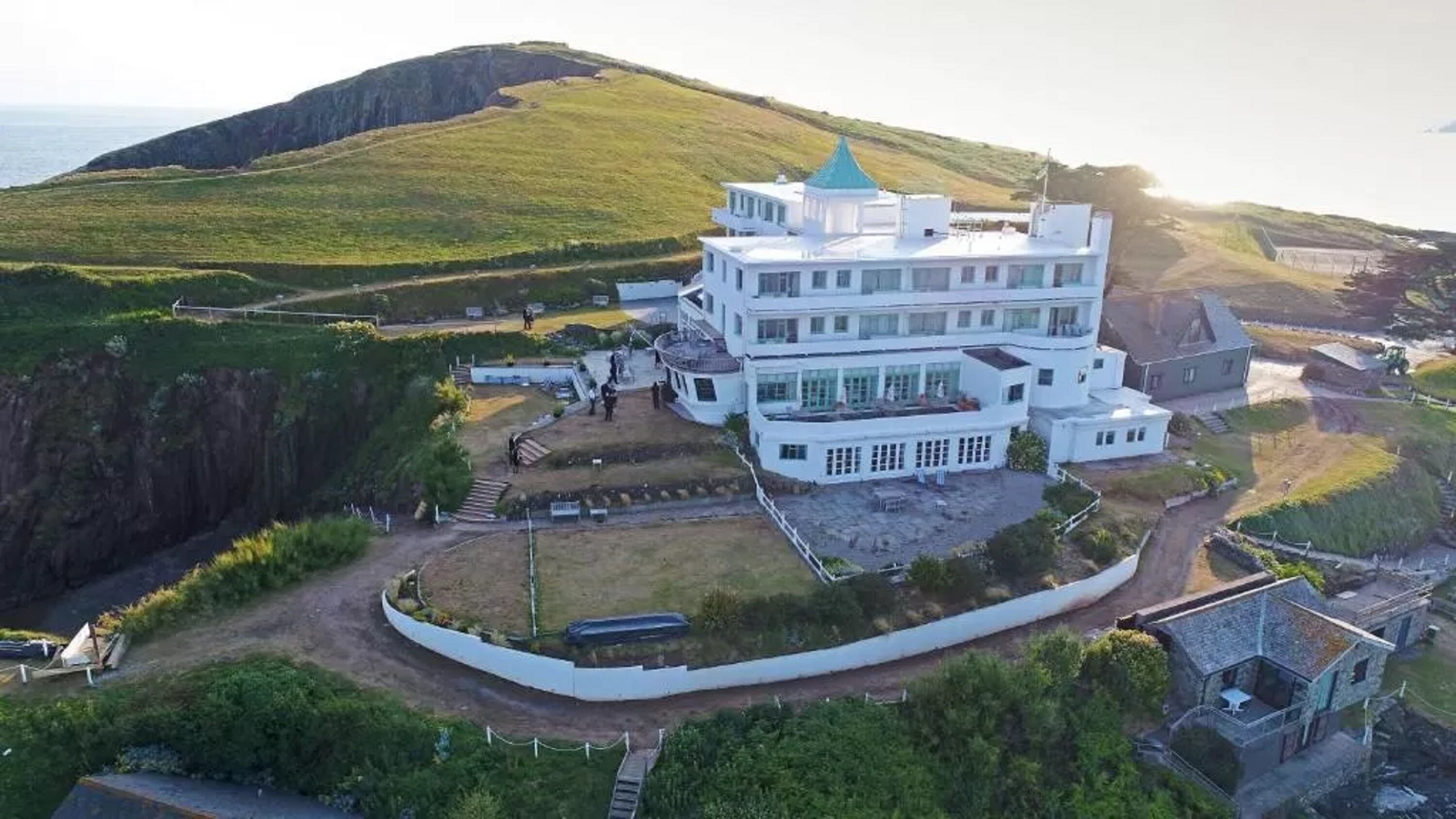Un grand bâtiment blanc à plusieurs étages, surmonté d’un toit végétalisé, entouré de collines verdoyantes et sous un ciel dégagé.
