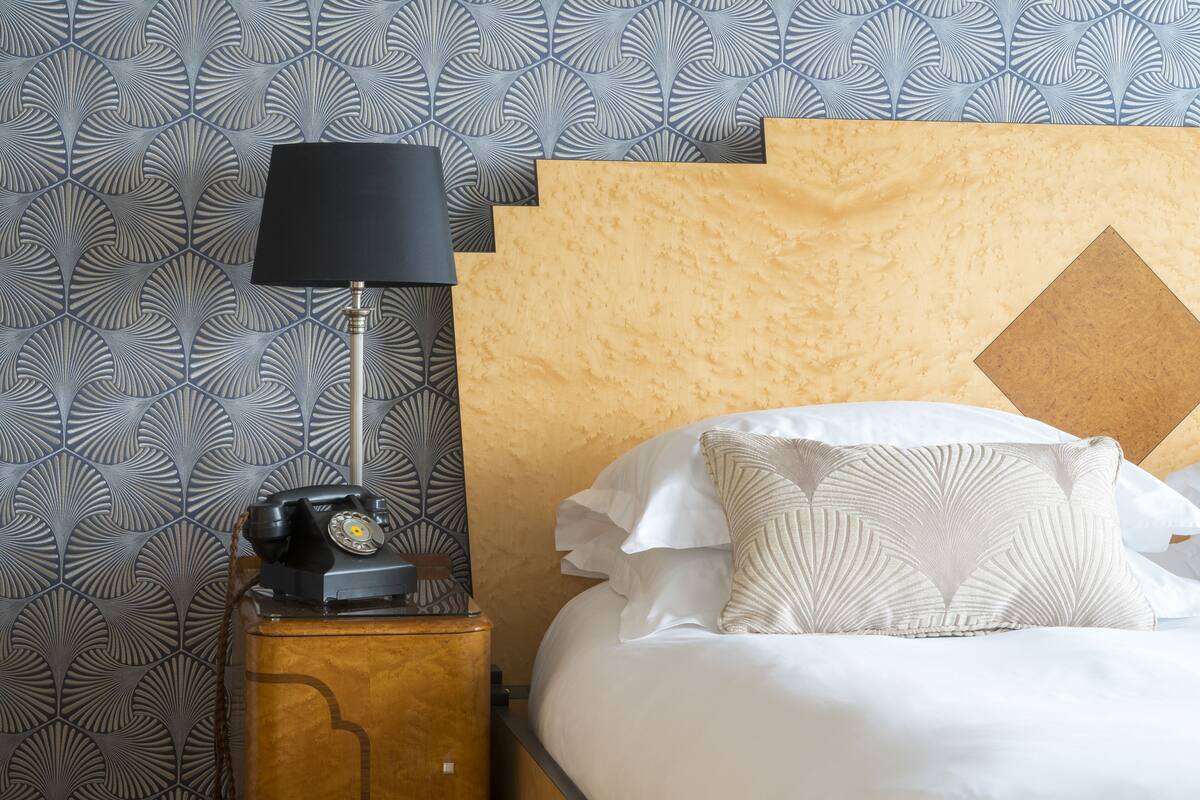 A neatly made bed with white linens and decorative pillows, a classic table lamp, and a vintage telephone.
