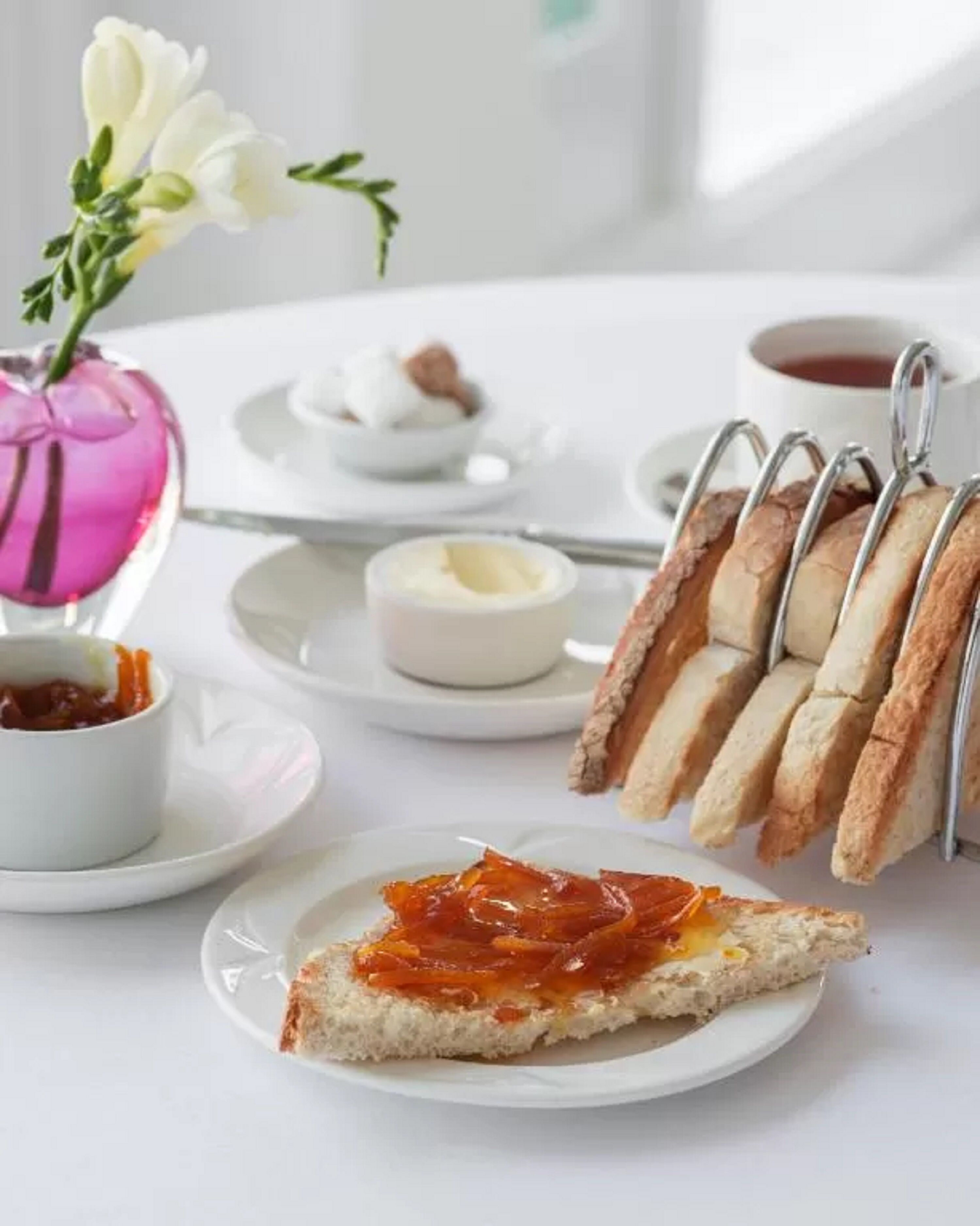 Una mesa dispuesta con tostadas y mermelada, un tazón pequeño de mermelada, una taza de té, un recipiente pequeño de mantequilla y un florero rosa con flores.
