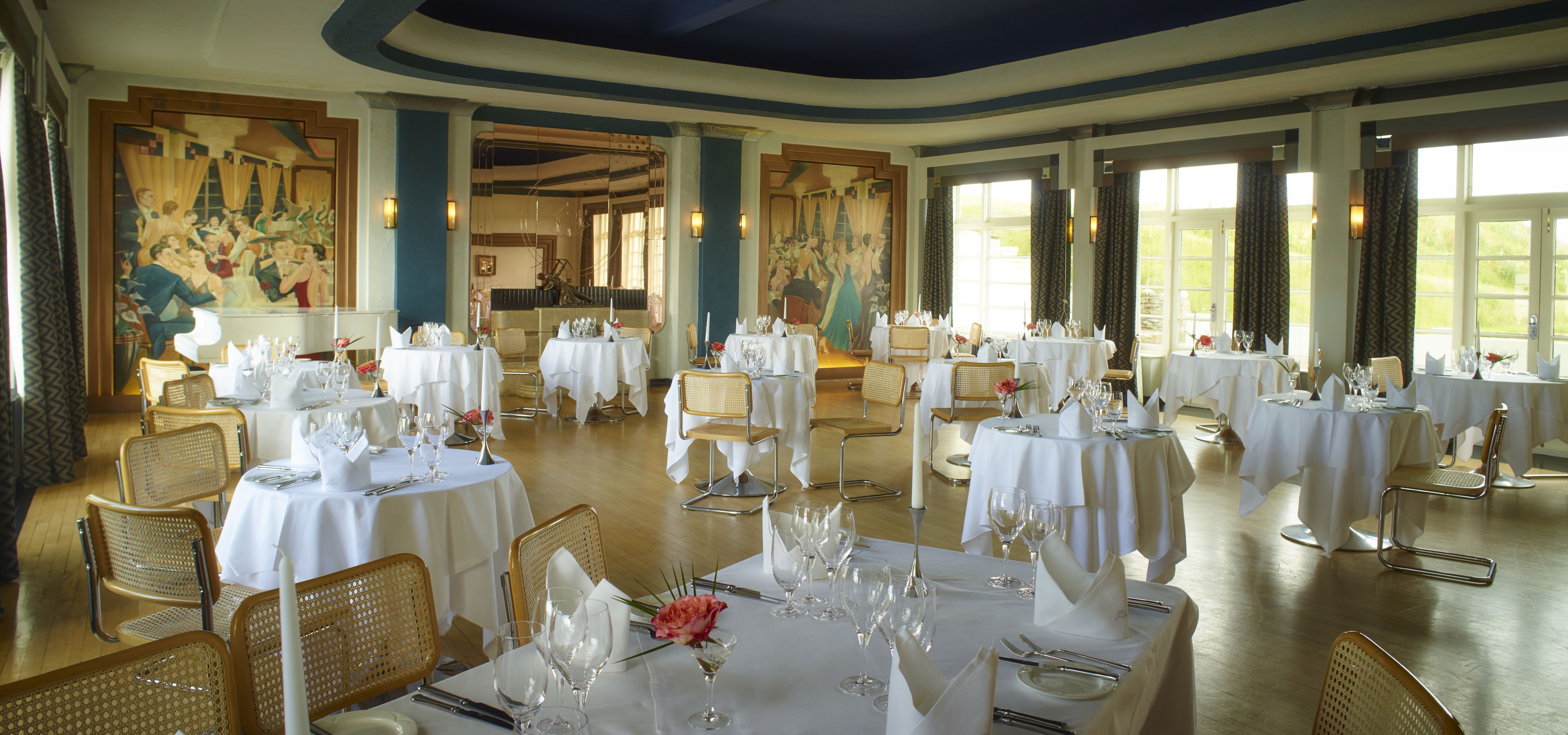 Un comedor con varias mesas dispuestas para un evento formal, con manteles blancos, cristalería y centros de mesa florales.