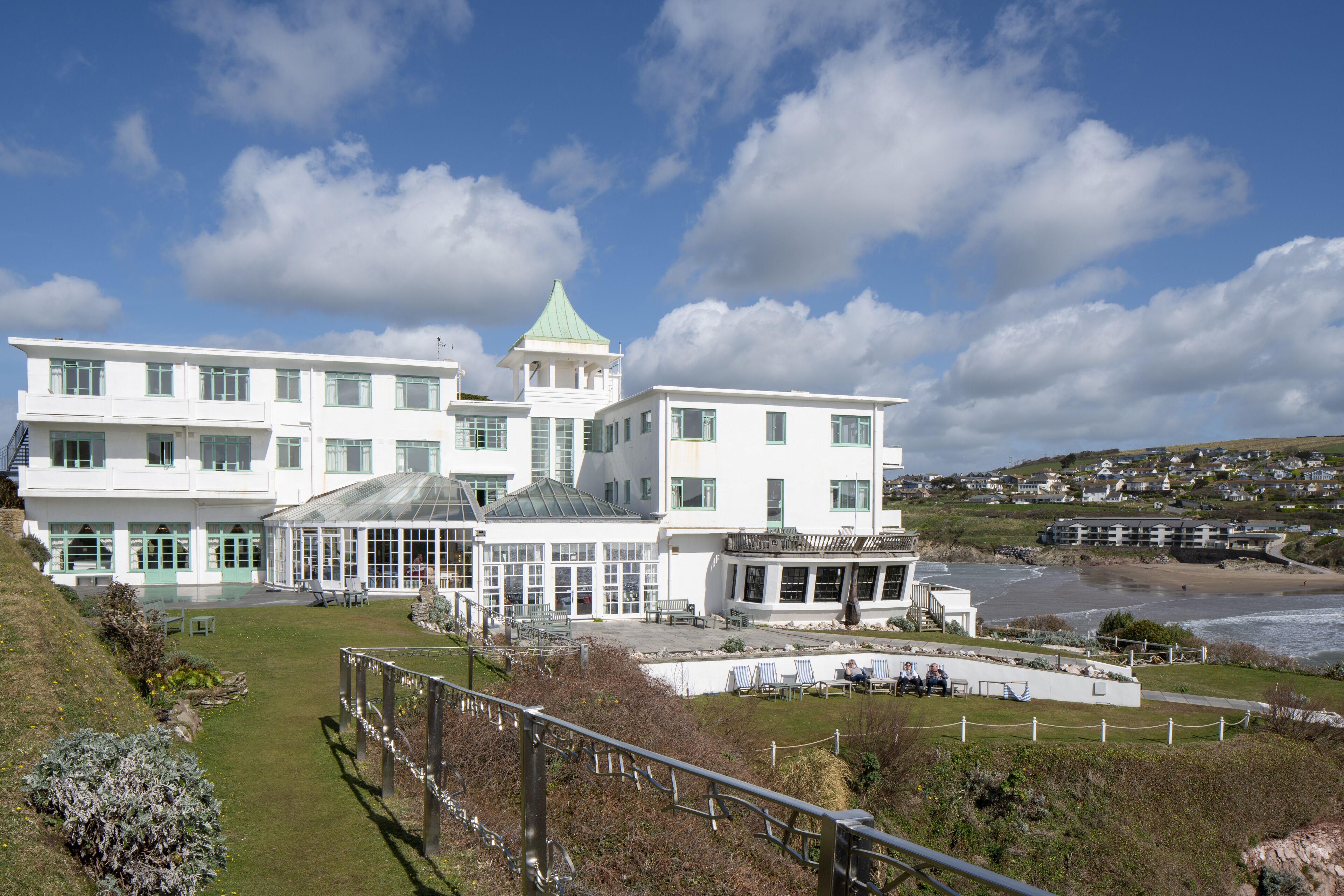A large white building with multiple windows and a green roof, situated near a beach with a view of the sea.