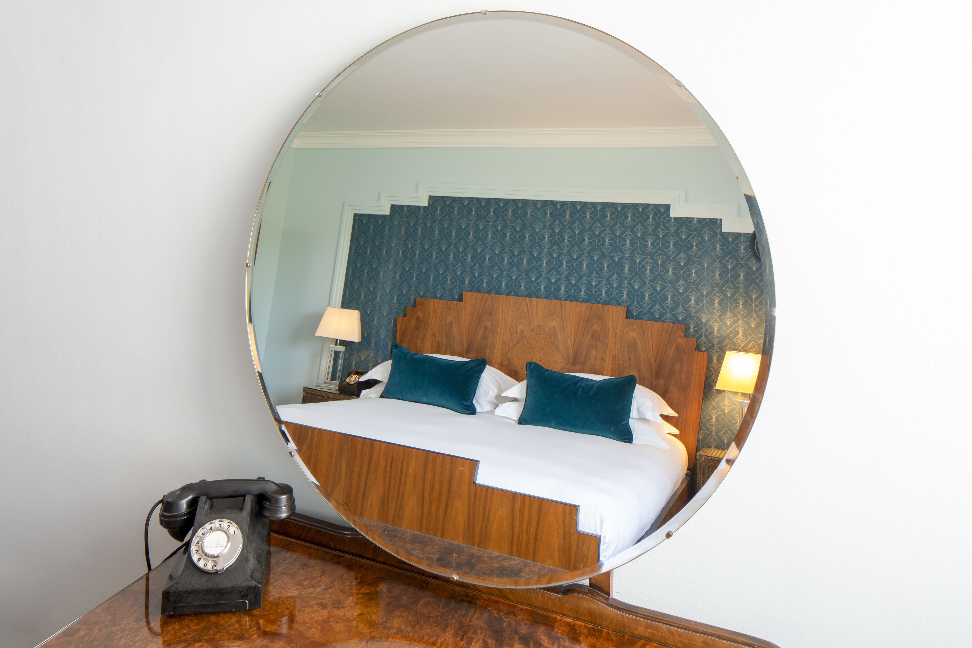 Een ronde spiegel weerspiegelt een bed met een houten hoofdbord, een nachtkastje met een lamp en een draaischijf telefoon op een houten oppervlak.