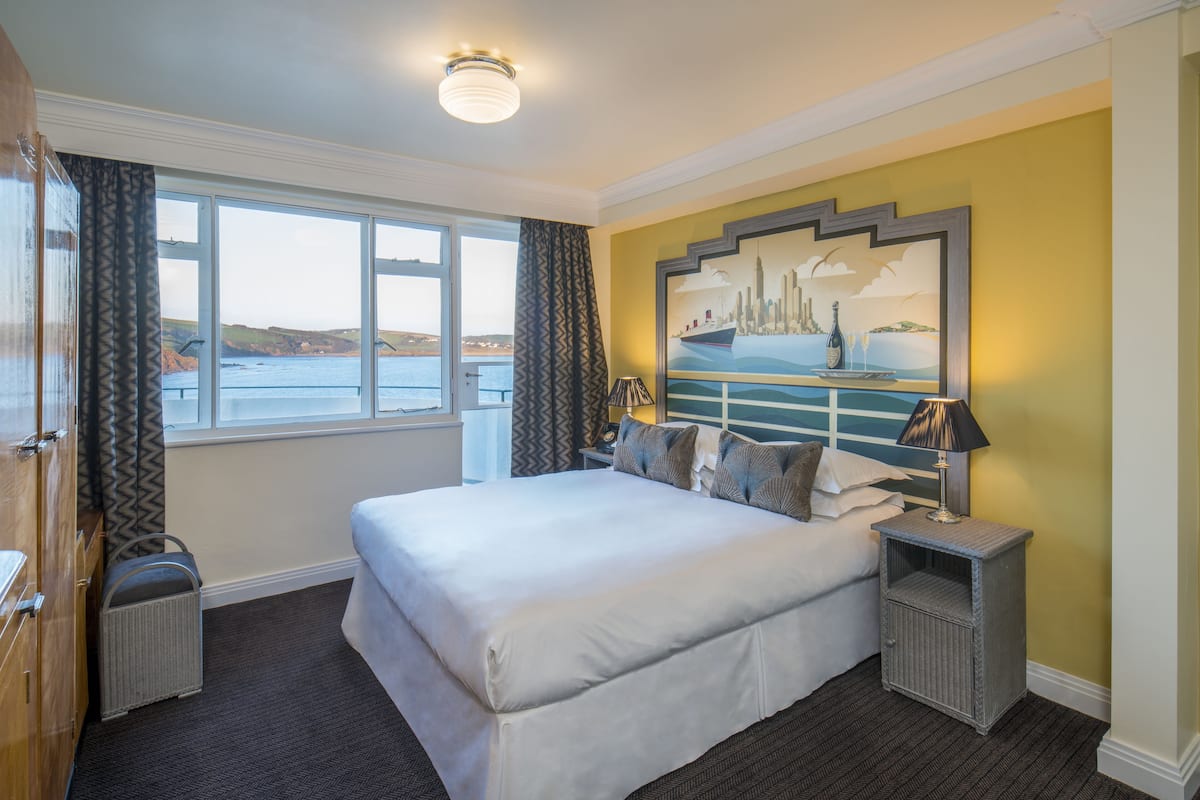 A hotel room with a large bed, a view of the sea, a painting on the wall, and a nightstand with a lamp.