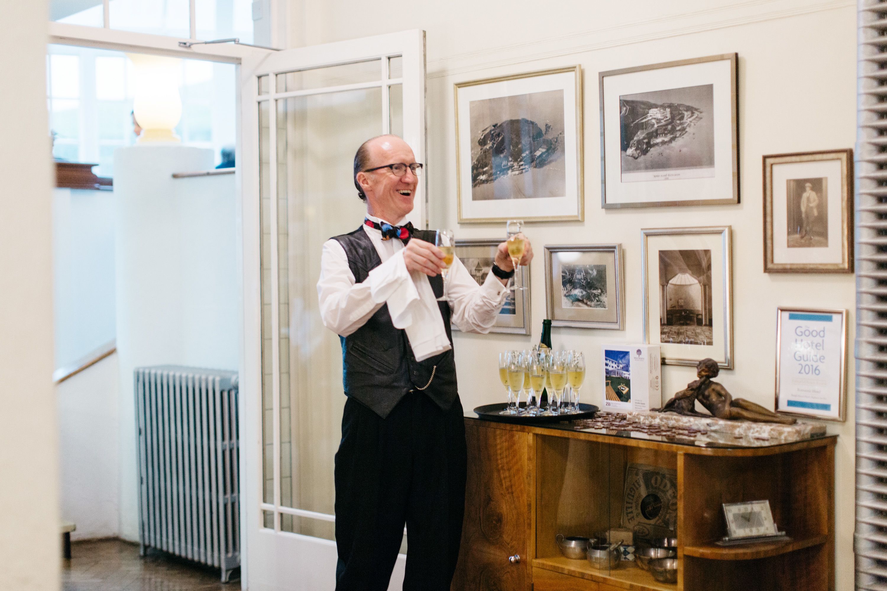 A man in a white shirt and black vest holding glasses, standing in a room with framed pictures on the wall and a wooden counter with glasses and bottles.