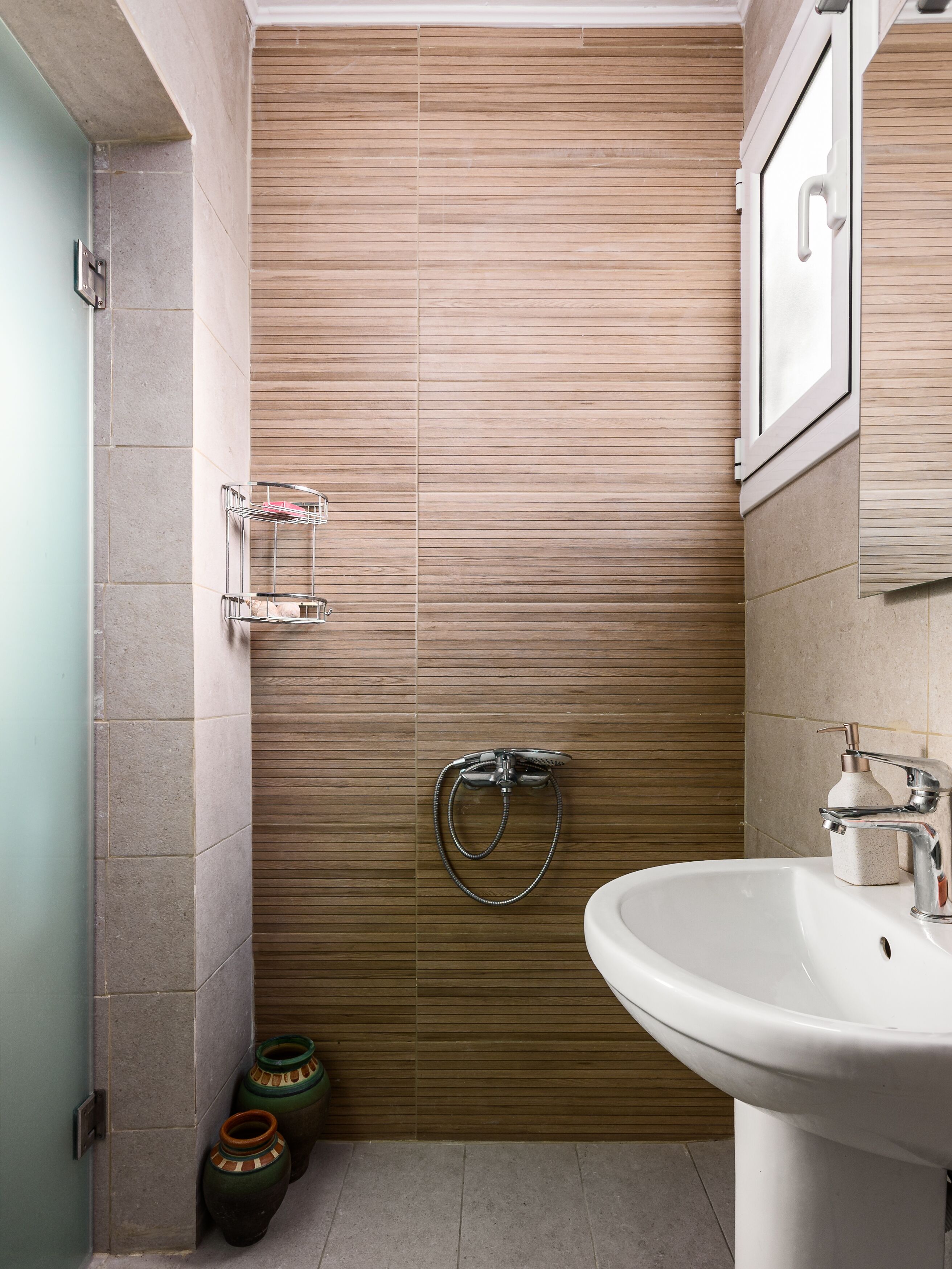 Une salle de bain avec un mur lambrissé en bois, une douche équipée d’un pommeau de douche à main, un lavabo avec un distributeur de savon et deux vases décoratifs.