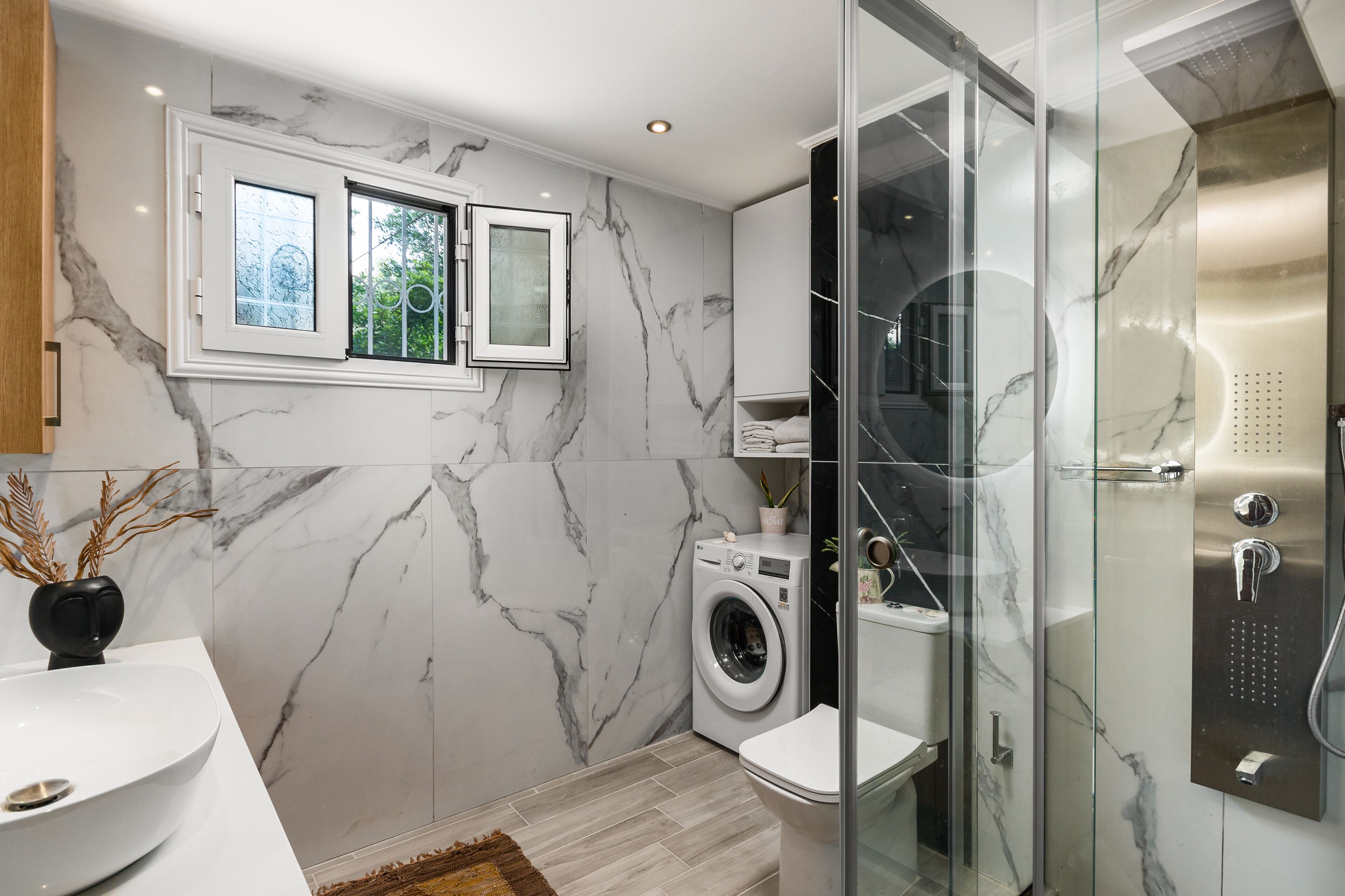 A modern bathroom with a marble wall, a washing machine, a glass-enclosed shower, and a window with a view of greenery.