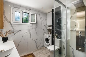 Een moderne badkamer met een marmeren wand, een wasmachine, een douche met glazen wanden en een raam met uitzicht op groen.