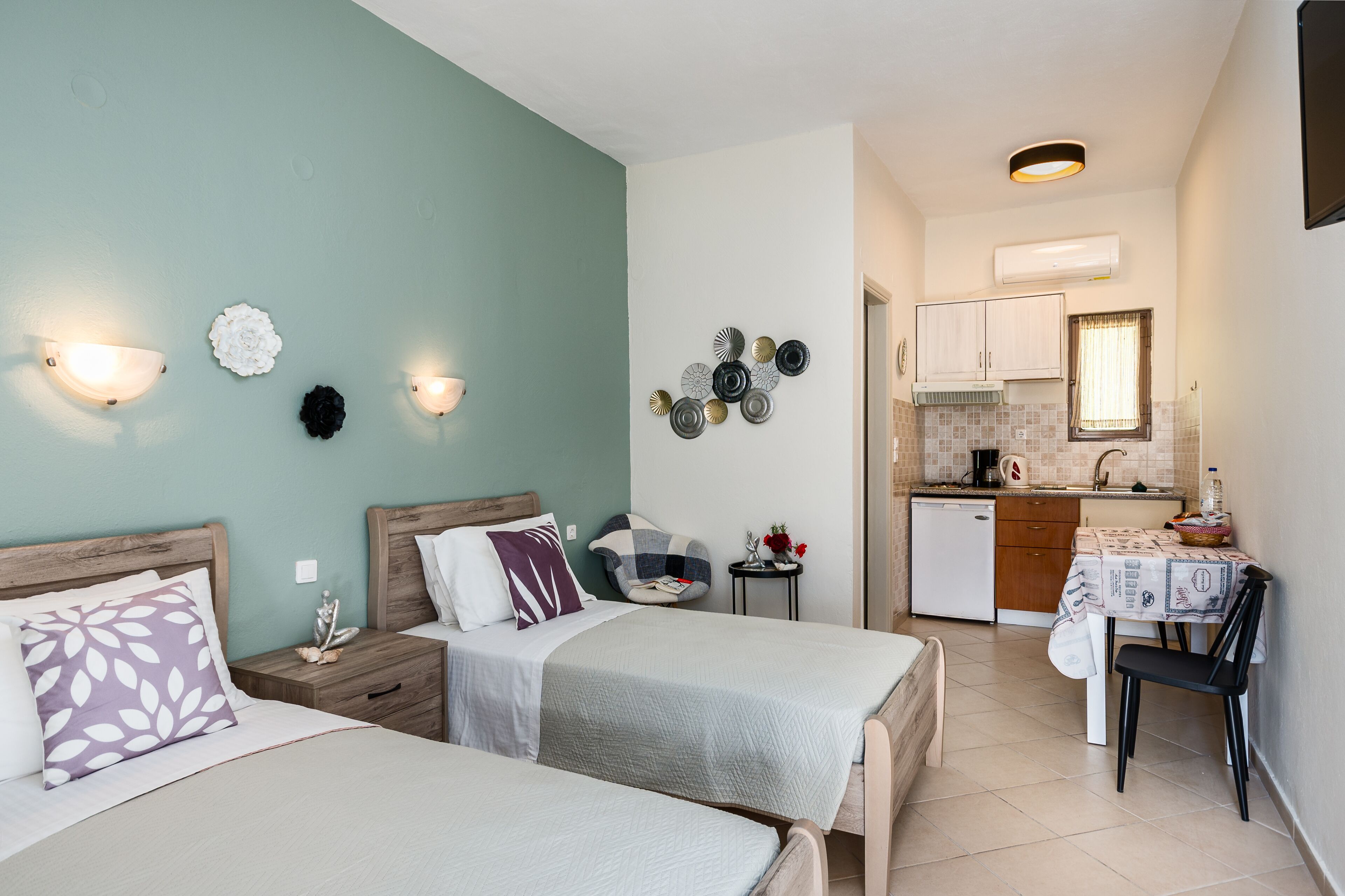 A bedroom with two beds, a small table, and a kitchenette in the background.