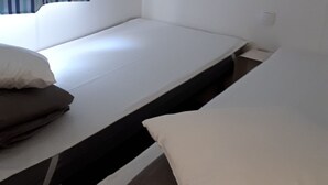Un lit simple avec un matelas blanc et deux oreillers, une petite étagère au-dessus et une fenêtre avec des rideaux à rayures.
