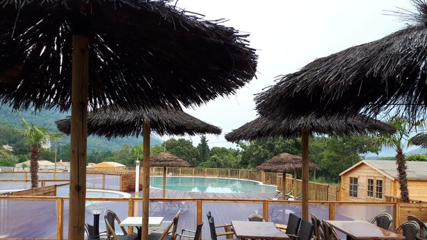 Une terrasse en bois avec des tables et des chaises, une piscine et des parasols en chaume.