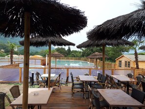 A wooden deck with tables and chairs, a pool, and thatched umbrellas.