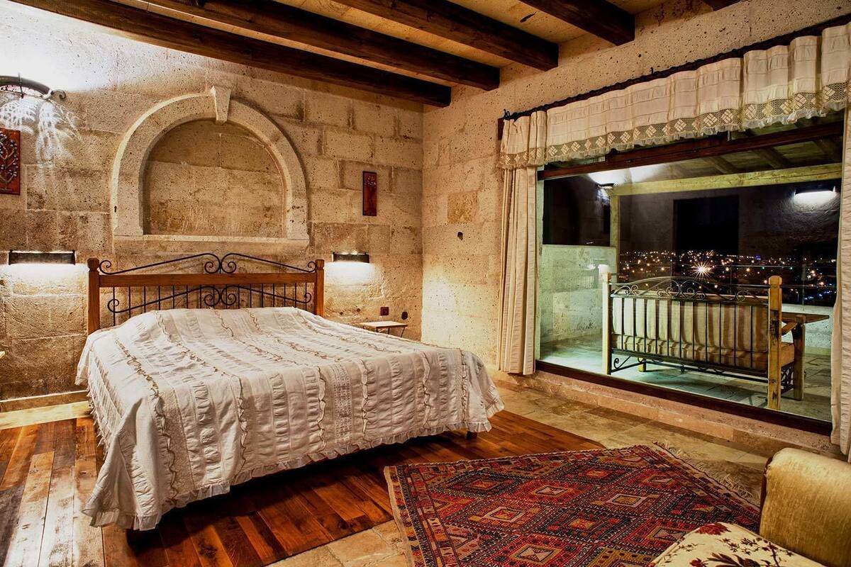 Quarto com cama de madeira, uma janela grande com varanda, paredes de pedra e teto rústico de madeira.