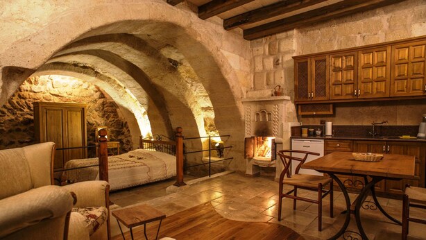 Interior rústico em pedra, com uma cama de madeira, uma mesa de jantar com cadeiras e uma área de cozinha com armários de madeira e lareira de pedra.